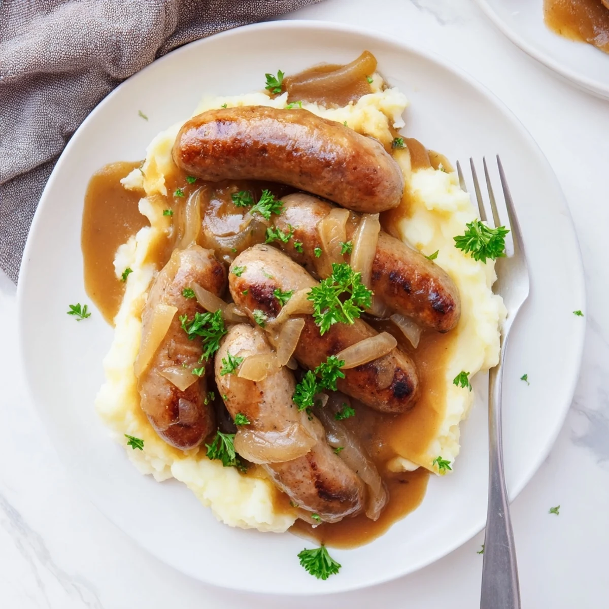 Close-up of golden brown Sausages in Onion Gravy in a cast iron skillet next to creamy mashed potatoes.